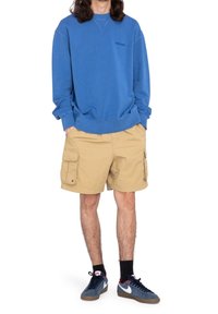 Felpa blu con polsini a coste, pantaloni cargo beige con più tasche e sneakers blu scuro con dettagli rossi e logo bianco.