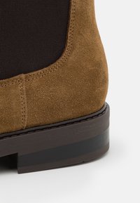 Hnědá semišová bota s černým elastickým panelem, s texturovaným povrchem a tmavě hnědou gumovou podrážkou. Prošívání podél okraje.