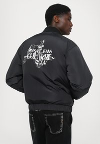 Vīrietis ģērbies melnā pūkainajā jaciņā ar baltu ziedu rakstu un uzrakstu "Versace Jeans Couture" mugurā, apvienojot to ar melnajām džinsām.