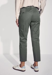 Grüne, zulaufende Hosen aus leichtem Stoff mit glatter Textur, ausgestattet mit Gesäßtaschen und getragen mit weißen und blauen Sneakers.
