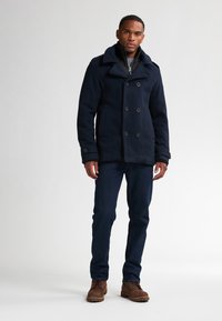 Pence di lana misto blu navy con chiusura a doppio petto, ampie rever, e tasche laterali, indossata sopra un maglione grigio e jeans scuri.