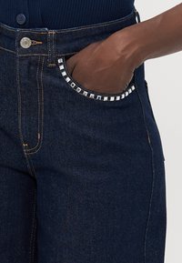 Mørke denimjeans med sølvfargede nagler på lommene, rett snitt og synlig gul søm langs sømmene.