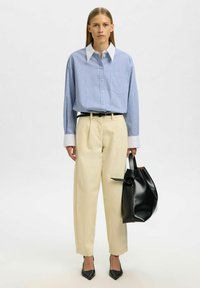 Chemise à rayures bleues avec col blanc, pantalon beige, sac à main en cuir noir et escarpins noirs pointus. Design simple et lignes épurées.