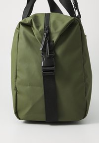 Sac en nylon vert avec un dessus zippé, une large ouverture et une sangle noire. Comprend une boucle en plastique et une surface texturée.