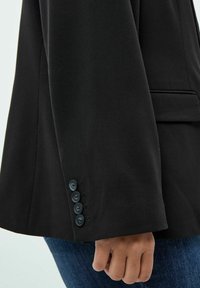 Manica di giacca nera con cinque bottoni neri, texture liscia, design sartoriale. Tasca visibile e forma leggermente aderente.