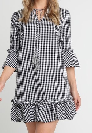 Czarna i biała sukienka w kratę z rękawami o długości trzy czwarte zakończonymi na dzwon, wiązaniem przy szyi z frędzlami oraz falbaną u dołu.