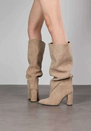 Jambes portant des bottes en daim beige jusqu'aux genoux, décontractées, avec des talons blocs, debout sur un sol gris lisse devant un fond clair uni.