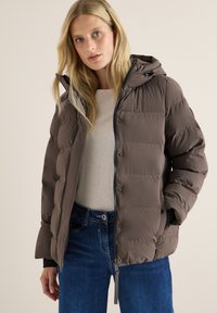 Veste matelassée marron avec capuche, présentant un devant zippé, des boutons-pression et un motif matelassé. Associée à un jean en denim clair.