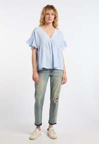 Lichtblauwe gestreepte blouse met ruches aan de mouwen en een V-hals, gecombineerd met vervaagde, gescheurde jeans en witte sneakers op een effen achtergrond.