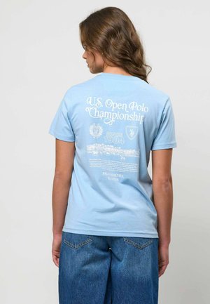 Pruunide juustega naine, kes kannab helesinist T-särki valge "U.S. Open Polo Championship" tekstiga ja graafikaga ning lahtisi siniseid teksapükse.