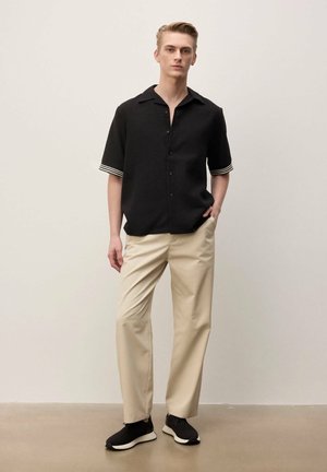 Jonge man staat en draagt een zwart overhemd met korte mouwen en gestreepte manchetten, beige broek en zwarte sneakers tegen een neutrale achtergrond.