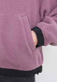 Sudadera de forro polar en púrpura claro con una superficie texturada, puños elásticos negros y detalles de bolsillo en la cintura.