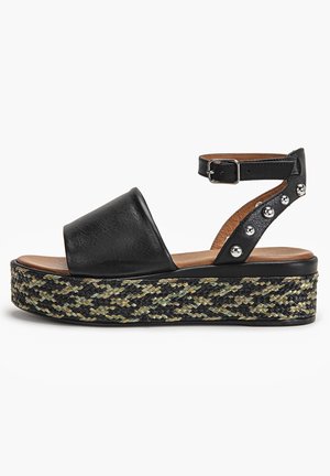 Czarne skórzane sandały mają szeroki przedni pasek, regulowany pasek wokół kostki z metalowymi ćwiekami oraz plecioną podeszwę na platformie w naturalnych odcieniach.