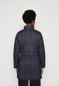 Abrigo acolchado de puffer en color azul marino, con cuello alto, mangas largas, cintura ceñida y costuras verticales sutiles, que exhibe una textura suave y brillante.