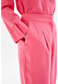 Combinaison rose vif cintrée avec manches longues, dotée d'une taille ceinturée, de boutons sur les manches et de poches latérales plissées.