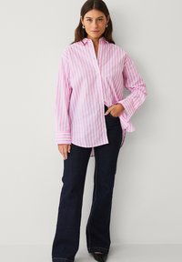 Chemise à rayures roses et blanches, boutonnée, à manches longues, associée à un jean évasé foncé. La chemise comporte un col et une coupe décontractée.