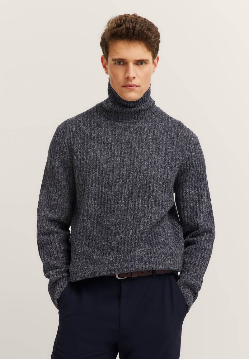 Dunkelgrauer Rollkragenpullover mit strukturiertem Rippenmuster, langen Ärmeln und lockerer Passform. Kombiniert mit dunklen Hosen, Hände in den Taschen.