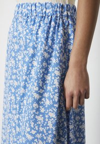 Modrá květinová sukně s bílými vzory, elastickým pasem a lehkým materiálem, zobrazující detail pasu a ruku, která drží sukni.