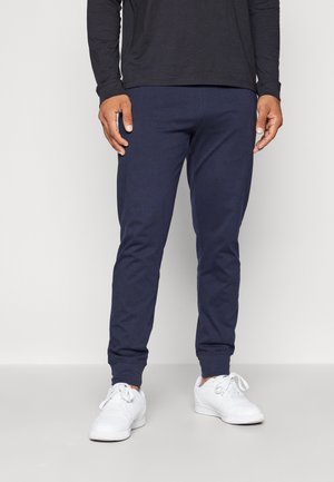 Homme portant un pantalon de jogging bleu marine, des baskets blanches et un t-shirt noir à manches longues, debout sur un sol gris clair.