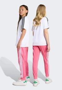 Dve dievčatá v bielych tričkách s čiernymi pruhmi a ružových teplákoch s bielymi pruhmi. Obidve majú na sebe topánky s zelenými akcentmi.