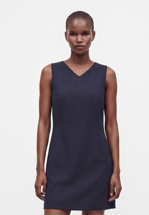 Femme aux cheveux courts portant une robe sans manches bleu marine foncé, longueur genou, se tenant devant un fond blanc uni.