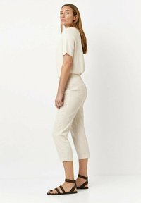 Hellbeige, cropped Hosen kombiniert mit einem passenden, kurzärmeligen Oberteil. Das Modell trägt braune Riemchensandalen. Glatte, leichte Stofftextur.