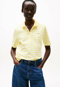 Camisa polo de rayas amarillas y blancas con un cuello clásico, mangas cortas y un pequeño logo cerca del pecho, combinada con pantalones cortos de mezclilla azul oscuro.