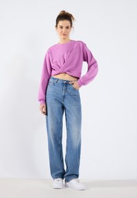 Rosa avskåret hettegenser med vridd frontdesign, kombinert med lyseblå vide jeans, komplementert av hvite sneakers. Minimal bakgrunn.