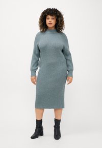 Ribbad långärmad klänning i mjuk teal med hög hals och rak silhuett, kombinerad med svarta ankelstövlar. Slät textur, längd till mitten av vaden.