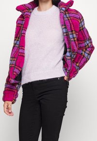 Veste en tartan rose et violet floue avec une fermeture éclair à l'avant, portée sur un pull en maille violet clair, associée à un pantalon noir. Les détails du tissu texturé sont visibles.