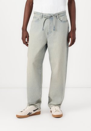 Lichtblauwe denim jeans met een ontspannen pasvorm, een verstelbare tailleband en een ontwerp met vijf zakken, gecombineerd met crèmekleurige sneakers met zwarte accenten.