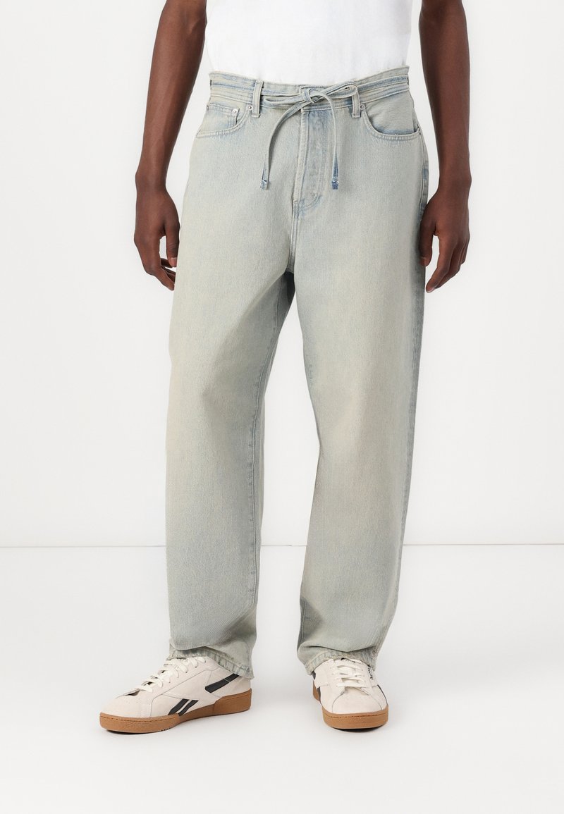 Lyseblå denimjeans med afslappet pasform, snøring i taljen og fem lommer, parret med cremefarvede sneakers med sorte accenter.