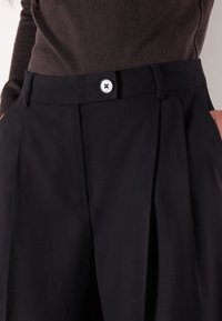 Sorte skræddersyede bukser med plisseringer, en glat tekstur og en enkelt sølvknap i taljen.