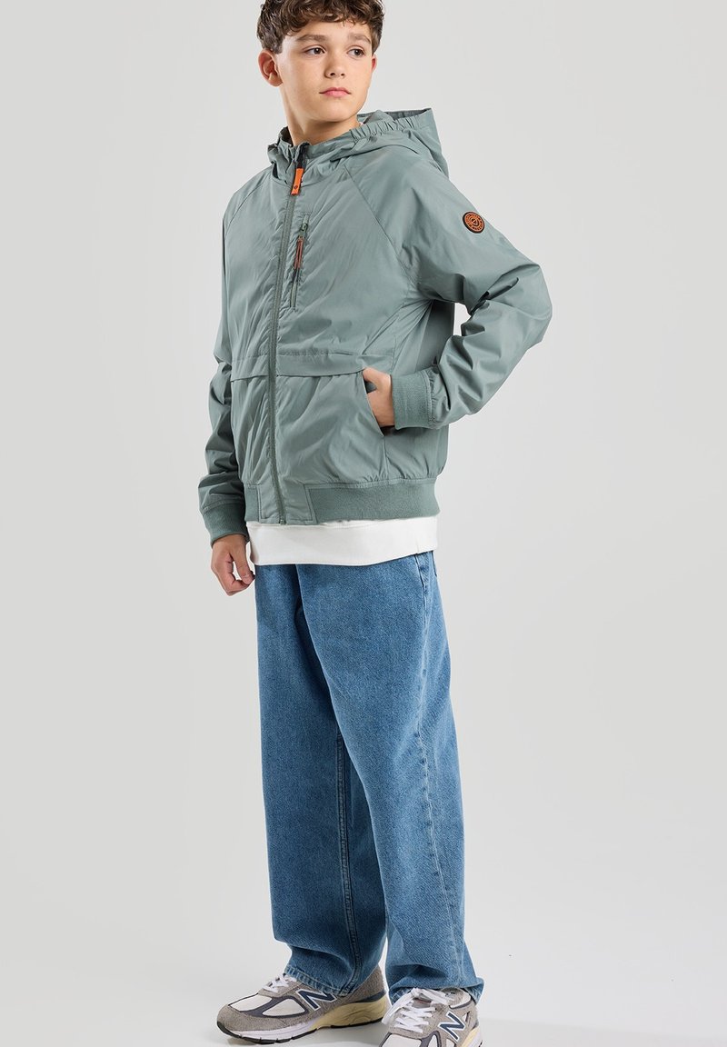 Vihreä vetoketjullinen takki hupulla, jossa on oranssi vetoketjun veto ja sivutaskut. Päällä löysät siniset farkut ja harmaat lenkkarit.