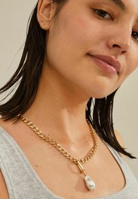Un collar de cadena de oro presenta eslabones grandes y un cierre, adornado con un colgante de perla barroca, sobre una blusa de color gris claro.
