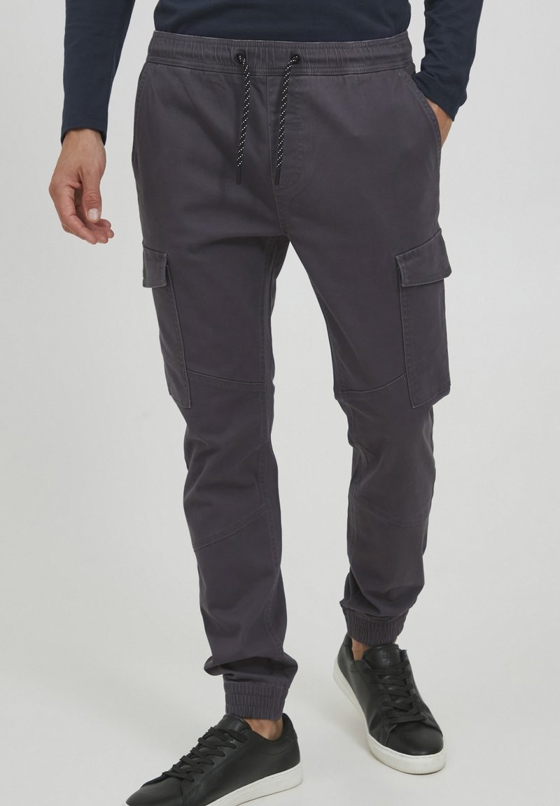 Homme portant un pantalon jogger cargo gris foncé avec poches latérales, chevilles élastiques, taille avec cordon de serrage, et des baskets noires à semelles blanches.