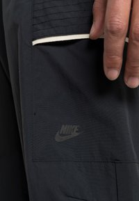Črn Nike material z gladko teksturo, s všitom stranskim žepom in majhnim črnim logotipom na spodnjem delu žepa.