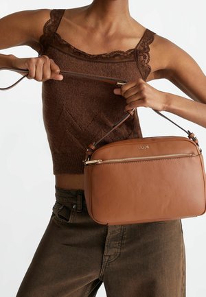 Personne portant un débardeur marron à dentelle et un pantalon foncé, tenant un sac bandoulière en cuir marron clair de la marque Liu Jo par sa sangle.