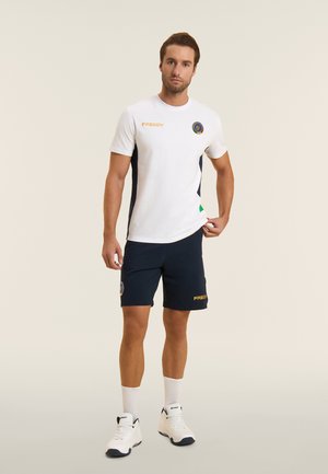 Uomo in piedi con t-shirt bianca e pantaloncini blu navy con loghi gialli "FREDDY", calzini bianchi e scarpe da ginnastica bianche su sfondo neutro.