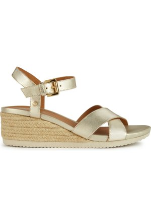 Sandal in metallo dorato con zeppa a cuneo, arricchita da fascette incrociate, fibbia alla caviglia e zeppa rivestita in juta. Parte superiore liscia e suola ammortizzata per maggiore comfort.