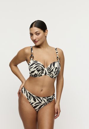 Vrouw met donker haar in een bikini met zebraprint, poseert met één hand op haar heup tegen een effen witte achtergrond.
