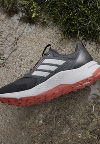 Chaussure de course noire et grise avec des bandes blanches et une semelle rose texturée sur une surface rugueuse en terre et herbe.
