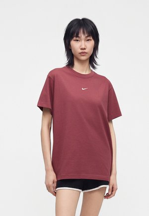 Jeune personne aux cheveux noirs portant un t-shirt Nike ample couleur bordeaux et un short noir avec bordure blanche, debout devant un fond blanc uni.