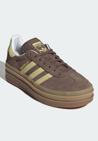 Brown Adidas Gazelle sneaker with beige stripes, thick sole, textured tongue, and white interior lining on a light background.