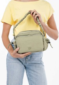 Borsa di stoffa verde con manico intrecciato, scomparti con zip e dettaglio del logo sulla parte frontale. Indossata su una maglietta gialla chiara e jeans di denim.