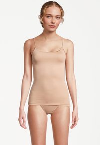 Woman with tied-back blonde hair wearing beige camisole and matching underwear, standing against plain white background.