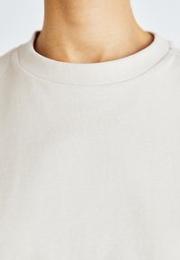 Světle béžové tričko s kulatým výstřihem a hladkou texturou. Tkanina je jednobarevná bez vzorů, což ukazuje na minimalistický design a volný střih.