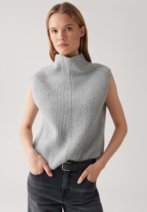 Vrouw in een mouwloze, grijze geribbelde truitje met col, gestopt in een zwarte spijkerbroek met een zwarte riem, staand met haar handen in de zakken.