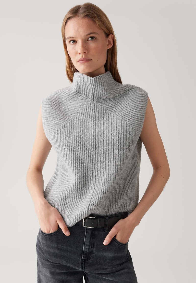 Frau in einem ärmellosen grauen, gerippten Rollkragenpullover, der in eine schwarze Jeans mit einem schwarzen Gürtel gesteckt ist, steht mit Händen in den Taschen.