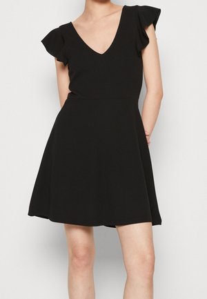 Woman wearing a black short dress with flutter sleeves and a V-neckline, standing against a plain light gray background.